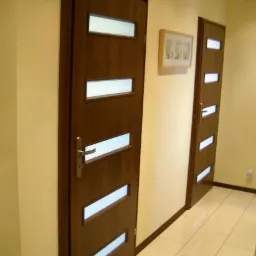 Wnętrze korytarza z dwiema drewnianymi drzwiami z przeszkleniami, widoczne płytki podłogowe w jasnym odcieniu i obrazek na ścianie.